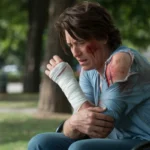 Injured person sitting on a park bench with bandages on their arm and shoulder, expressing pain after a dog bite or accident.